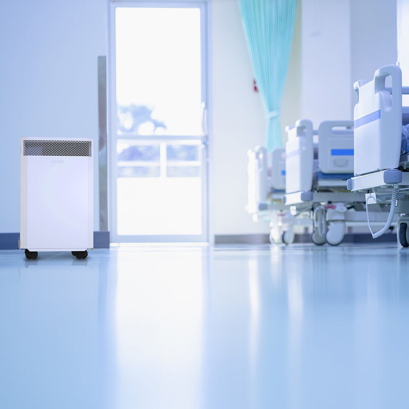 Load image into Gallery viewer, A hospital ward with the foot end of three beds visible. An INOVA air purifier has been placed by the entry doorway and is cleaning the air, reducing the risk of airborne viruses.